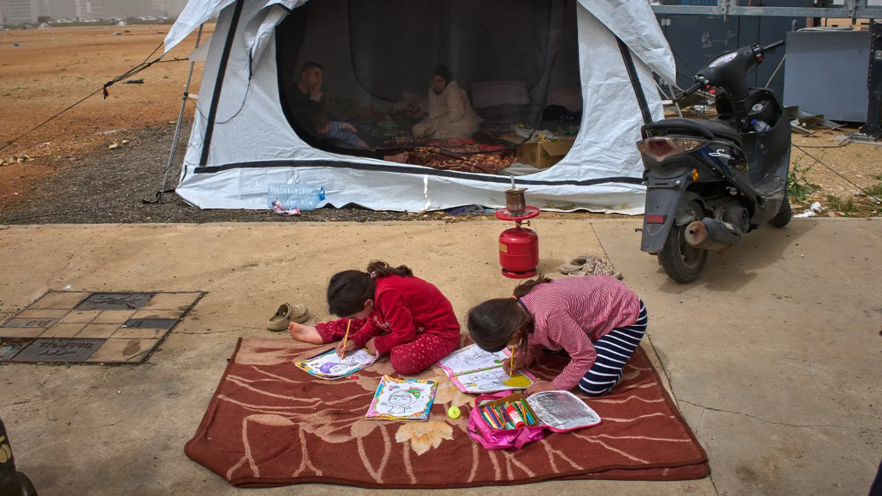 Tamara and her sister Amal color pictures on the floor as their parents, Sara and Ahmed, who fled their village of Khiyam in southern Lebanon due to Israeli bombardment, sit inside a tent used as a shelter in Beirut, Lebanon, Friday, April 3, 2026. (AP Photo/Emilio Morenatti)