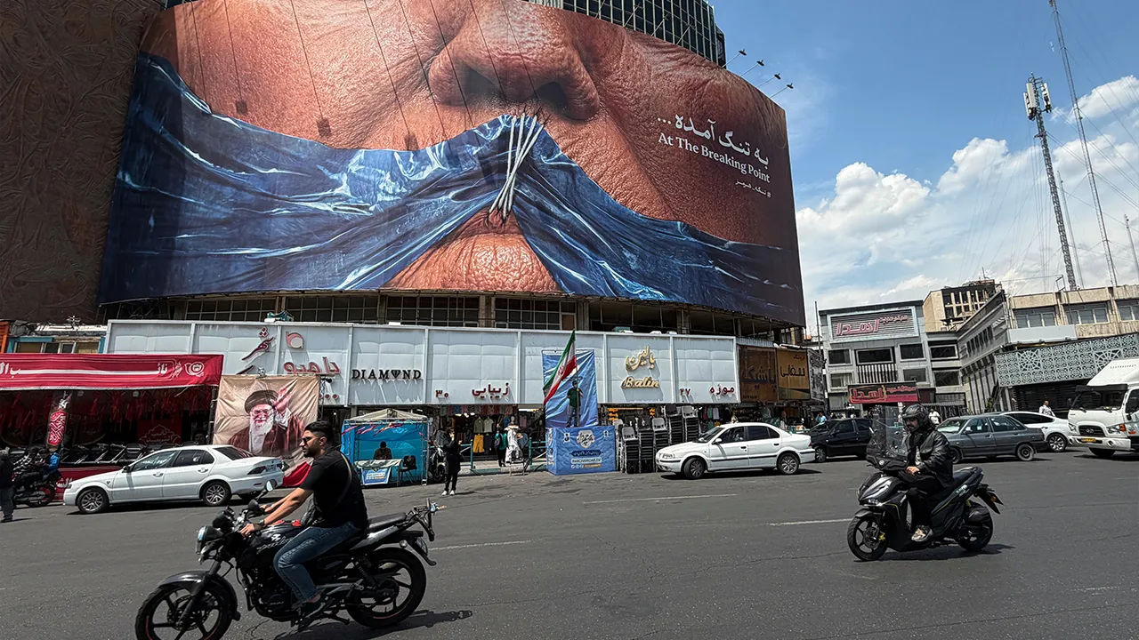 Vehicles drive past a billboard with graphic showing Strait of Hormuz and sewn lips of U.S. President Donald Trump in a square in downtown Tehran, Iran, Saturday, May 2, 2026. (AP Photo/Vahid Salemi)