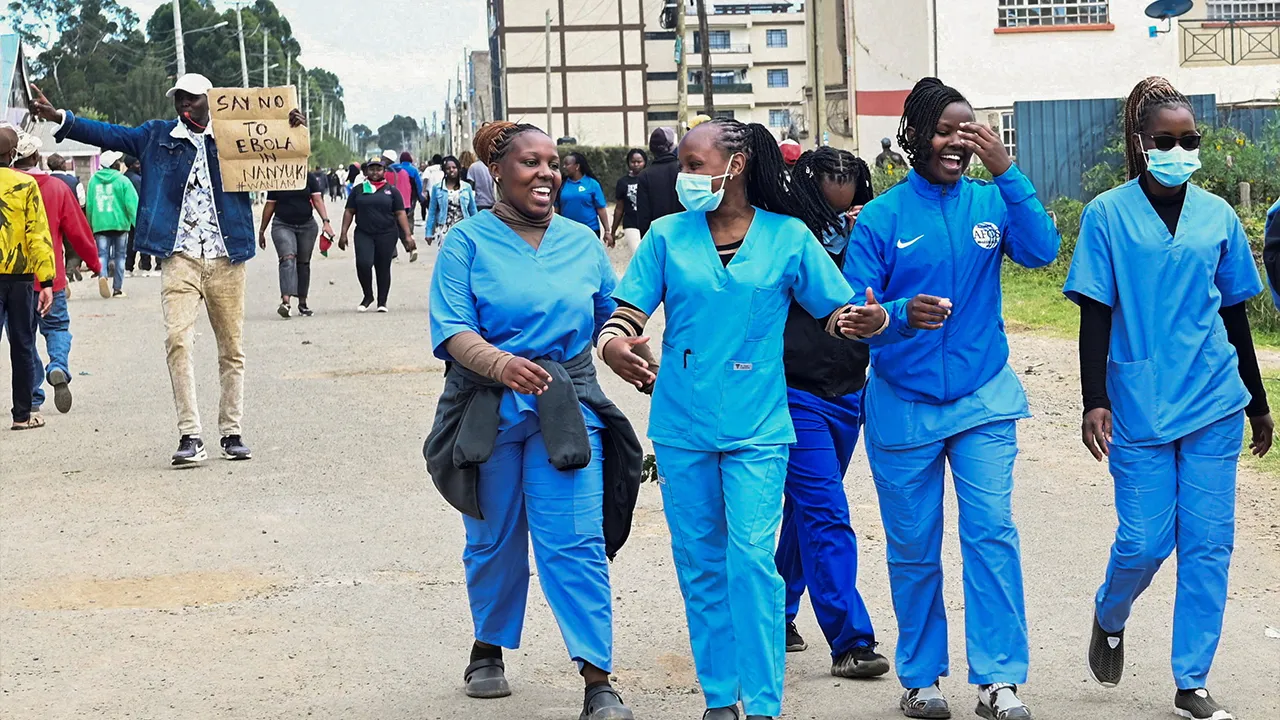 Women dressed in scrubs walk along the street as demonstrators participate during a protest against a U.S.-backed Ebola quarantine plan on the establishment of a 50-bed facility at a Kenyan air force base that was intended to host Americans exposed to Ebola, in Nanyuki town, in Laikipia County, Kenya June 1, 2026. REUTERS/John Muchucha REFILE - QUALITY REPEAT