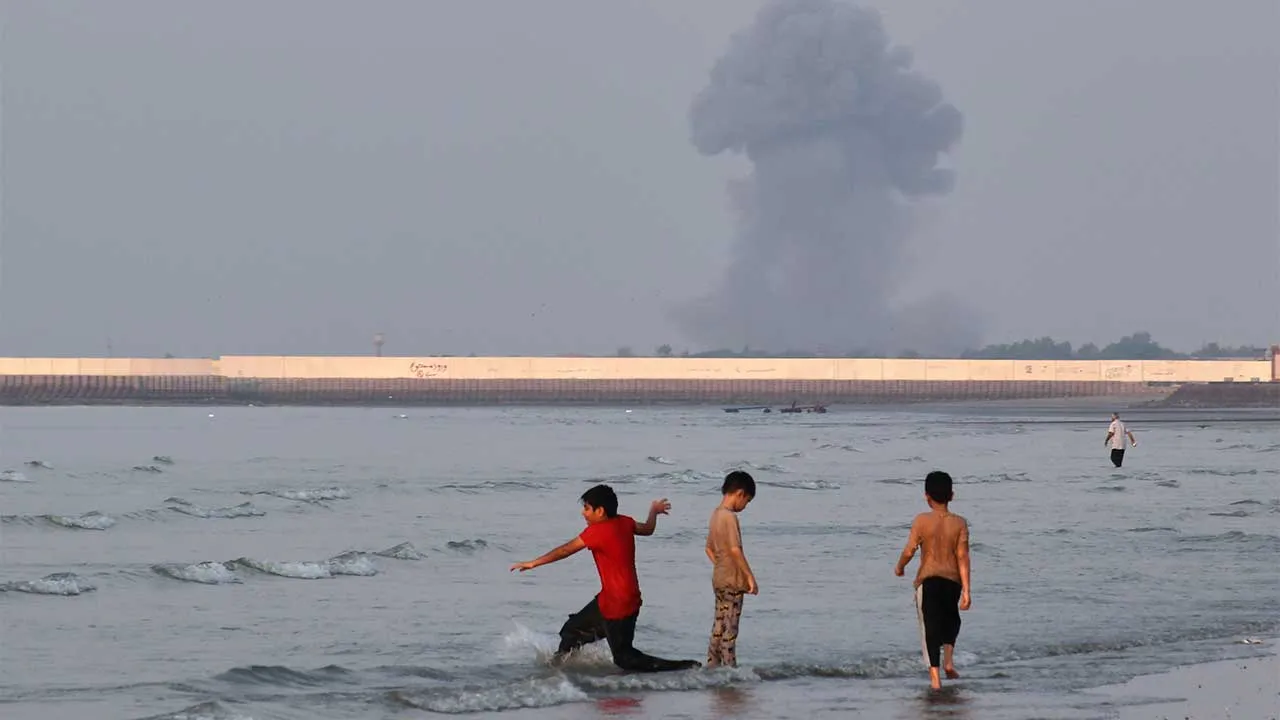 Three boys play in the shallow waters of the Strait of Hormuz, as a plume of smoke rises from an explosion in the background, off Bandar Abbas, Iran, Monday, July 13, 2026. (Razieh Poudat/ISNA via AP)