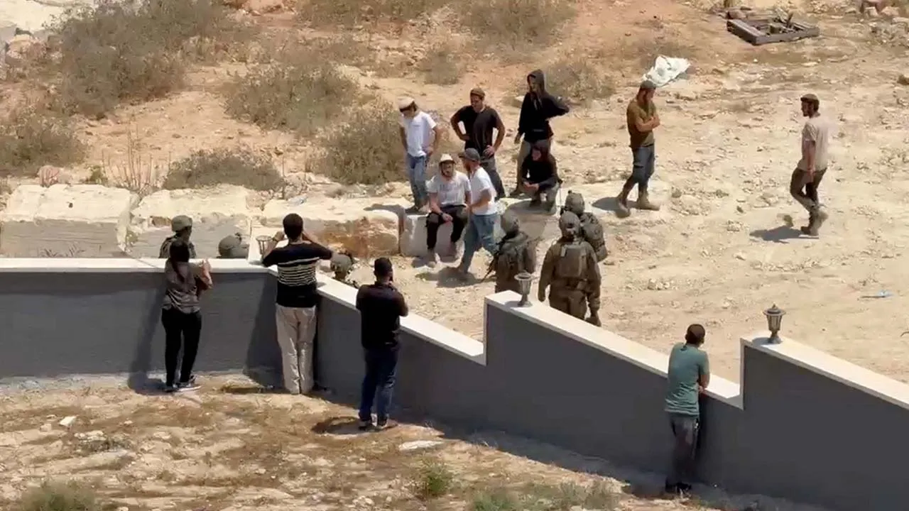 Palestinians use their phones as Israeli settlers and military personnel stand outside a Palestinian home in Qusra, in the Israeli-occupied West Bank, August 15, 2026, in this screen grab obtained from a video. VOBR/via REUTERS THIS IMAGE HAS BEEN SUPPLIED BY A THIRD PARTY. MANDATORY CREDIT. Verification: - Buildings, walls, gate, road layout and terrain matched satellite imagery and other videos verified by Reuters. - Date verified by Reuters talking to the source.  Preview Download Preview  XML
