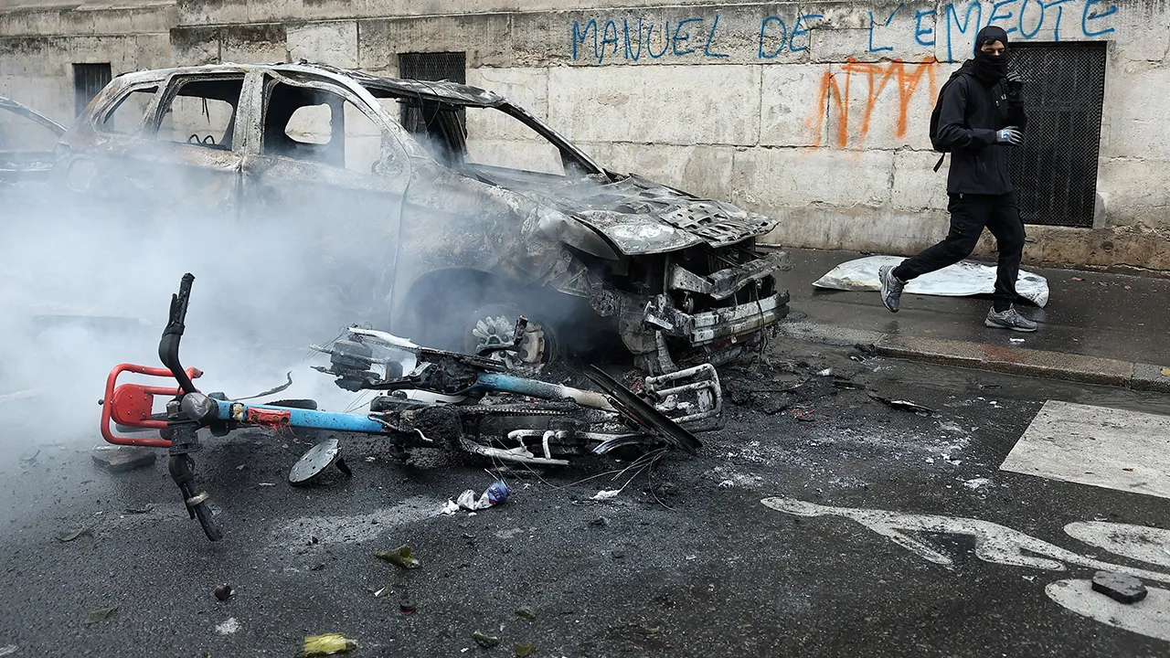 FILE PHOTO: A masked protester walks past a car and a bicycle burned during a demonstration against the cost of living and rising fuel prices near Place de la Republique in Paris as part of nationwide protests called by the "Yellow Vests" movement, joined by French high school and university students, teachers and parents demanding better school conditions and government action, as well as labour unions, workers, farmers and retirees, France, October 17, 2026. The slogan on the wall reads "Riot manual". REU