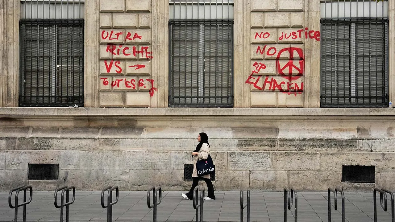 A woman walks past a building with graffiti that reads "The ultra-rich against everyone" and "No justice, no peace. RIP El Hacen" after the weekend protests over the cost of living turned into rioting and looting across France, in Paris, Wednesday, Oct. 21, 2026. (AP Photo/Teresa Suarez)