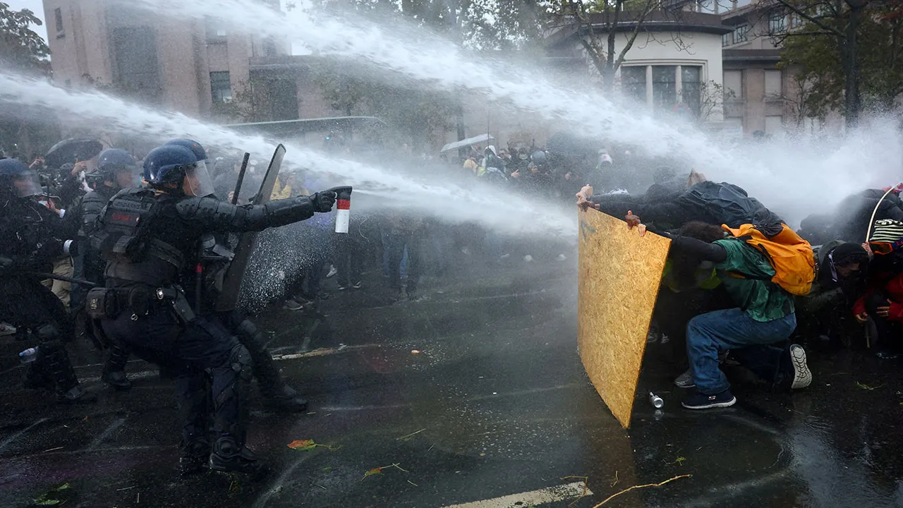 Protesters take cover behind a plywood board as French riot police and gendarmes use a water canon amid clashes during an authorised demonstration organised by labour unions in Paris as part of nationwide protests called by the "Yellow Vests" movement joined by French high school and university students against the cost of living and rising fuel prices, in France, October 24, 2026. REUTERS/Hannah McKay